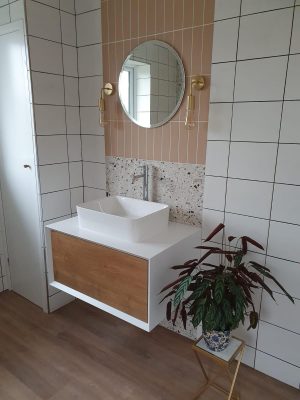 Straight swap bathroom door view of vanity unit complete, comprising of a white and wood effect cupboard and sink mounted on top with mono-mixer tap. 2 types of tiles and mirror with wall mounted pendant lighting.