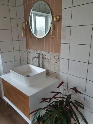 Straight swap bathroom door view of vanity unit complete, comprising of a white and wood effect cupboard and sink mounted on top with mono-mixer tap. 2 types of tiles and mirror with wall mounted pendant lighting.