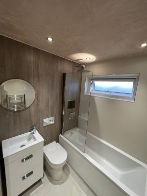 compact bathroom suite with round mirror, floating two drawer vanity unit and compact toilet with hidden cistern and rain shower head over bath with glass shower screen