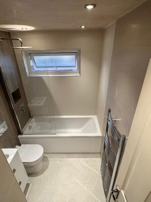 top view of compact bathroom suite with round mirror, floating two drawer vanity unit and compact toilet with hidden cistern and rain shower head over bath with glass shower screen
