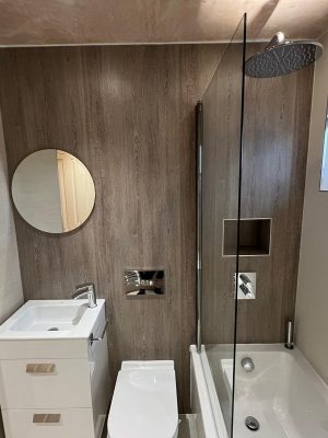 compact bathroom suite side wall with round mirror, floating two drawer vanity unit and compact toilet with hidden cistern and rain shower head over bath with glass shower screen