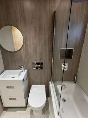 compact bathroom suite side wall with round mirror, floating two drawer vanity unit and compact toilet with hidden cistern and rain shower head over bath with glass shower screen
